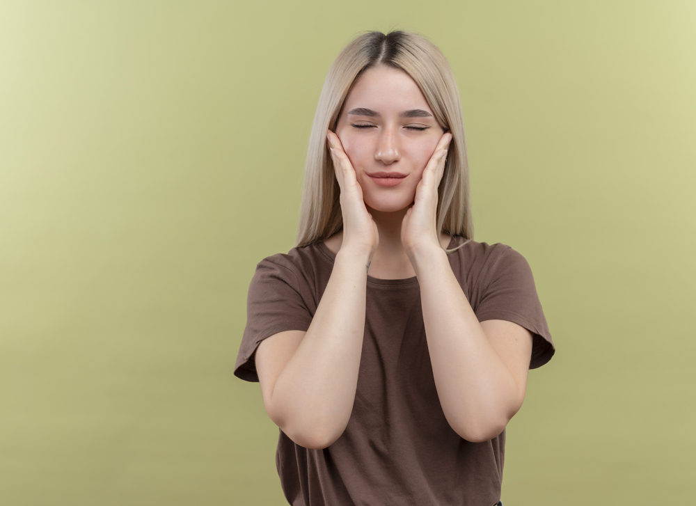 peaceful young blonde girl putting hands on cheeks with closed eyes on isolated green background with copy space