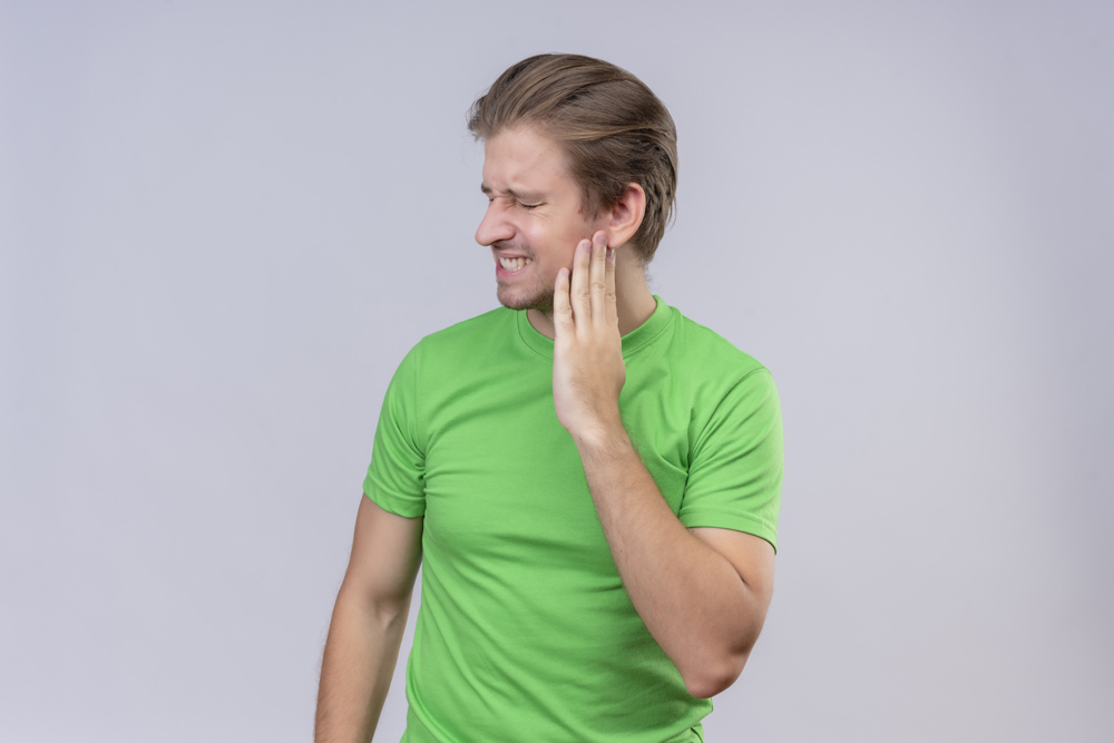 young handsome man wearing green t-shirt looking unwell touching his cheek having toothache standing over white background
